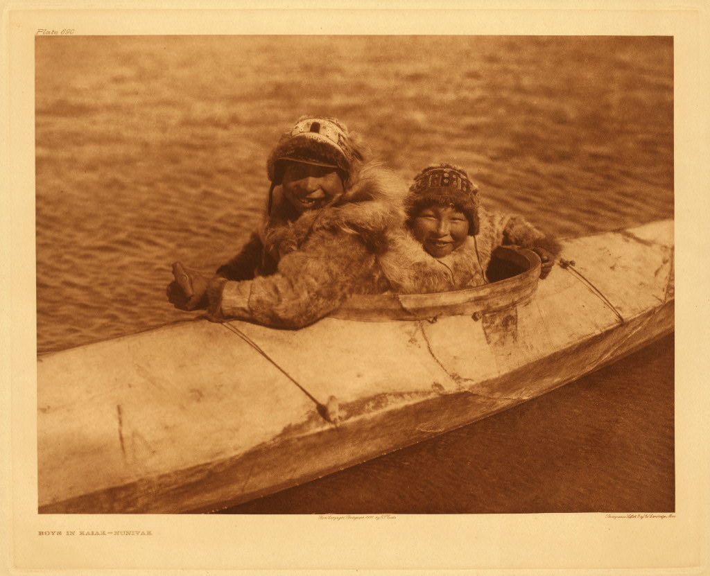 Photo of two males wearing fur sitting in well of large kayak