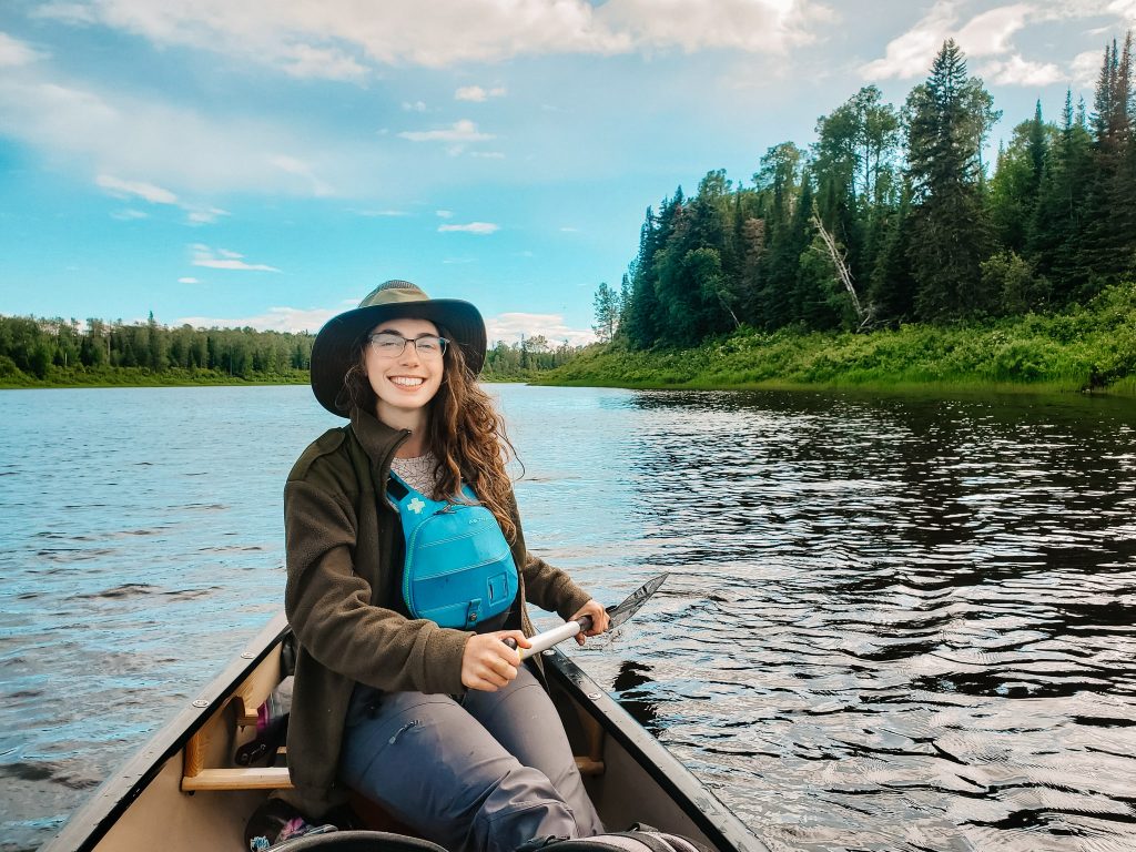 Woman sitting in a canoe wearing a life jacket for canoeing , big hat and holding a paddle on a canoe camping trip.