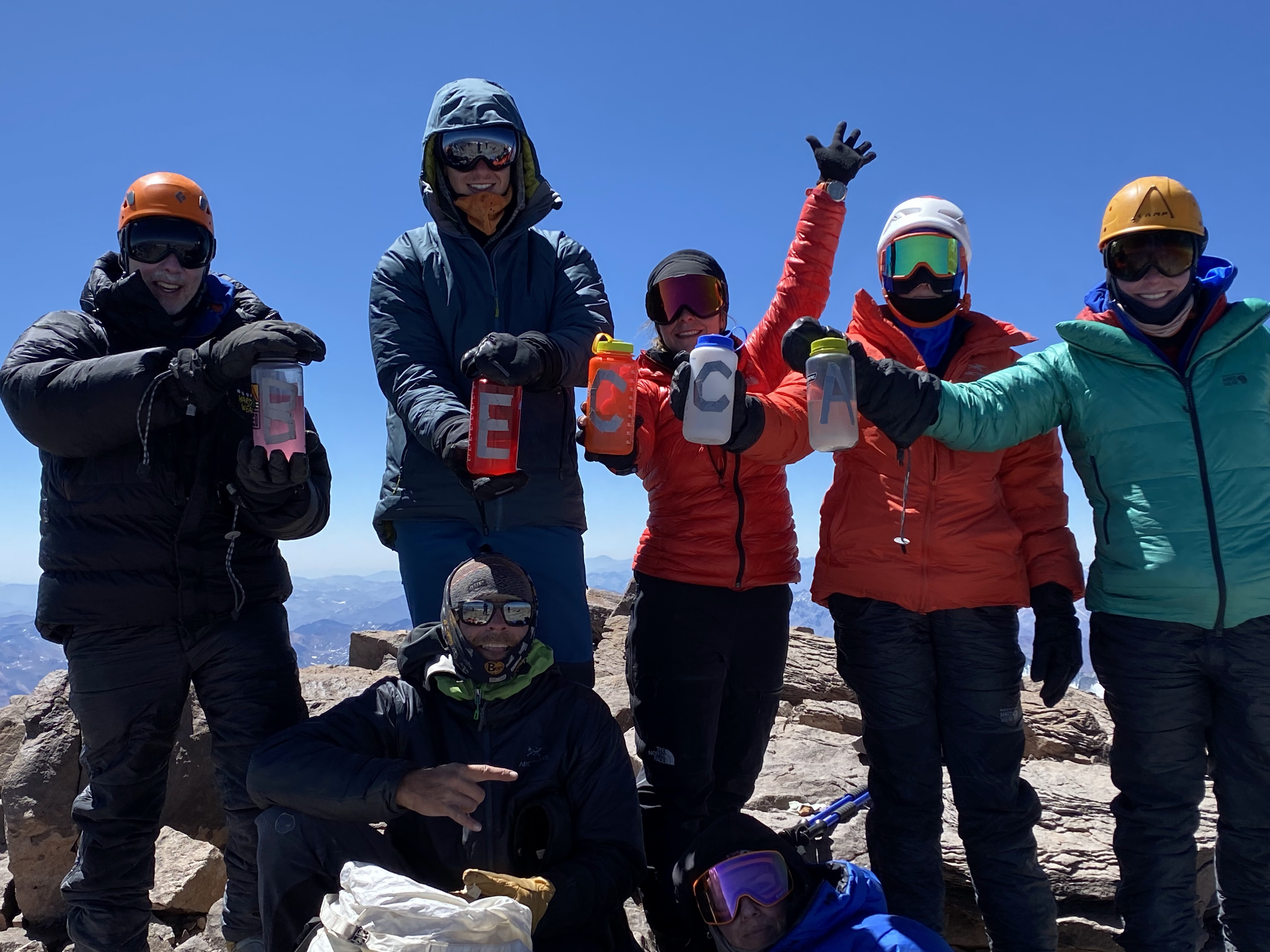 Шесть человек стоят на вершине Аконкагуа и держат бутылки с водой, на которых написаны буквы: БЕККА.