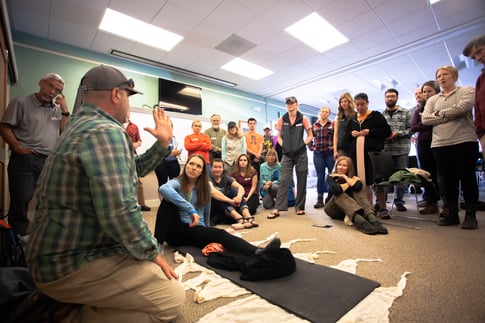 Группа студентов стоит вокруг человека, лежащего на полу.  Мужчина на переднем плане руками показывает, как выглядит позвоночник.